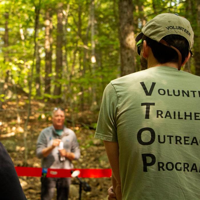 Man stands with back to camera wearing green shirt with text reading "Volunteer Trailhead Outreach Program"