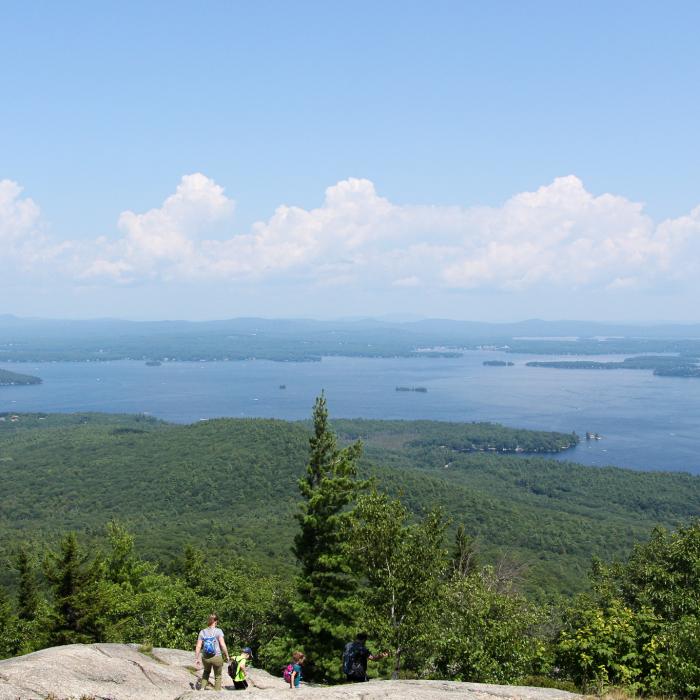 Two people hike down a rocky face with a large lake in the distance