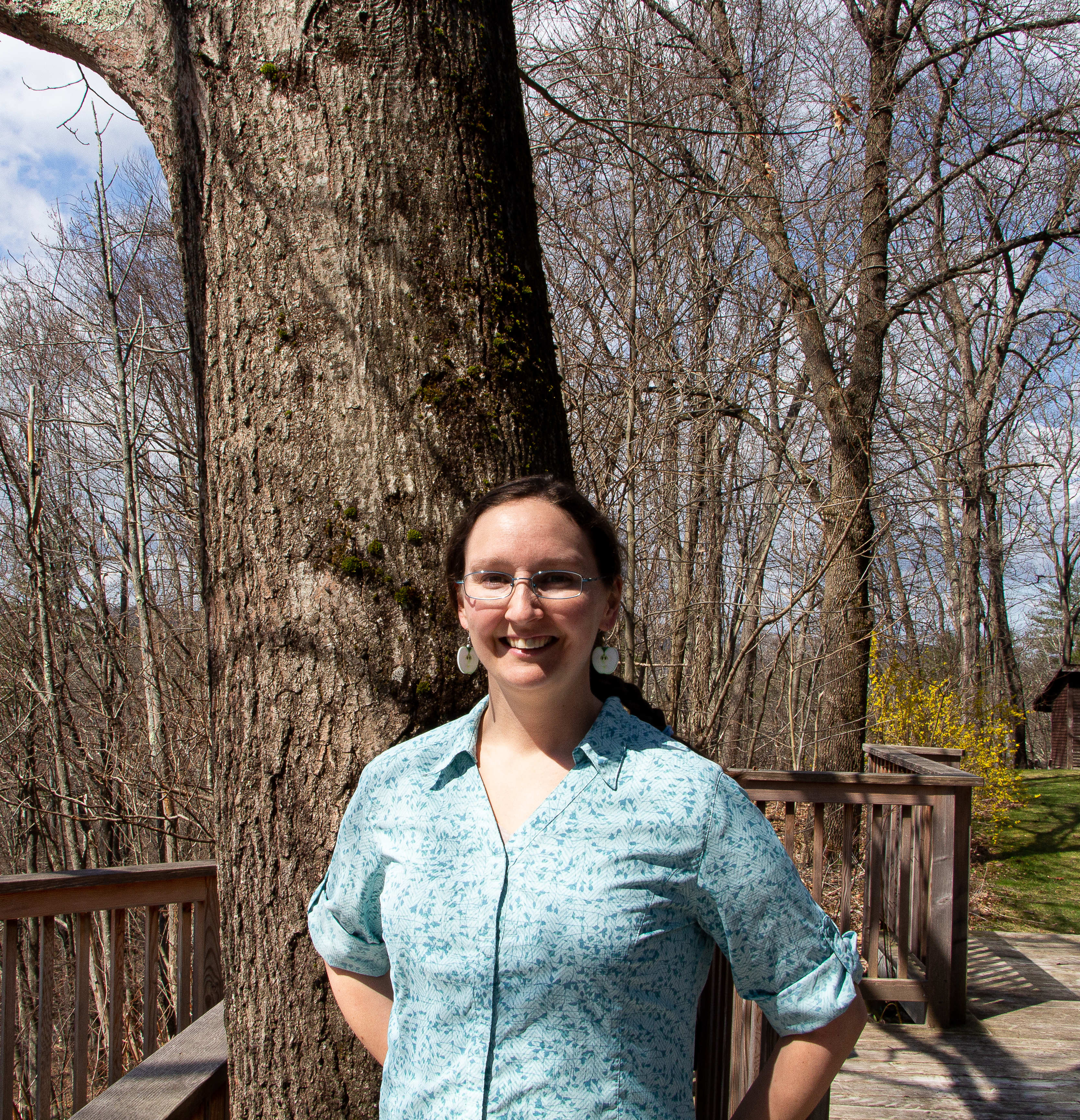 Young woman in a blue button up shirt stands in front of a tree