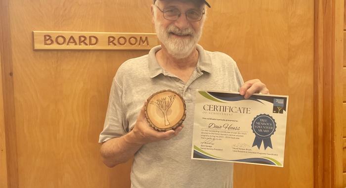 A man in a baseball hat holds up a paper certificate and a tree cookie award. 