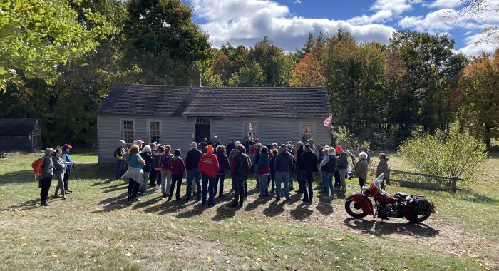 A group of people stand in front of a small grey house in a field on a sunny fall day. 