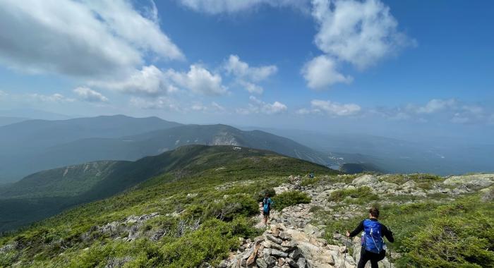 A woman with a blue backpack hikes over a rocky alpine trail. 