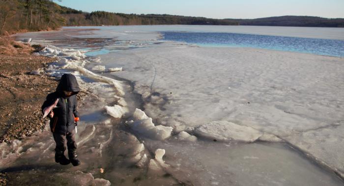 A boy in snow boots stands near an icy lake holding a pinwheel.