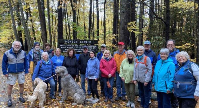 Colorfully clad group of older hikers pose at Monson Center sign