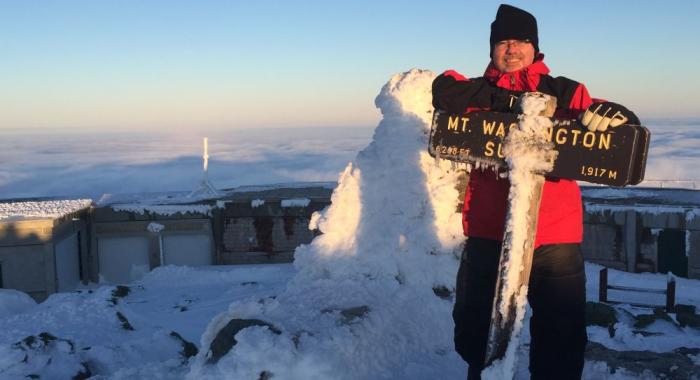 Dan Szczesny in red parka poses at rime ice crusted summit sign on Mt Washington in winter