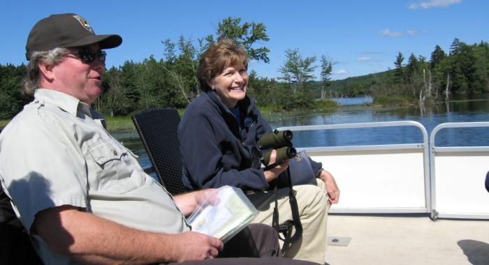 Senator Jeanne Shaheen smiling with binoculars sitting on boat with USFW Staff on Lake Umbagog on a clear blue sky day.