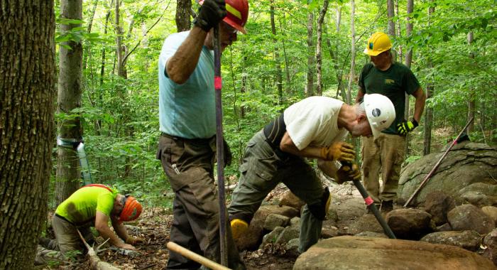 Three men wearing hardhats work to move a large rock. 