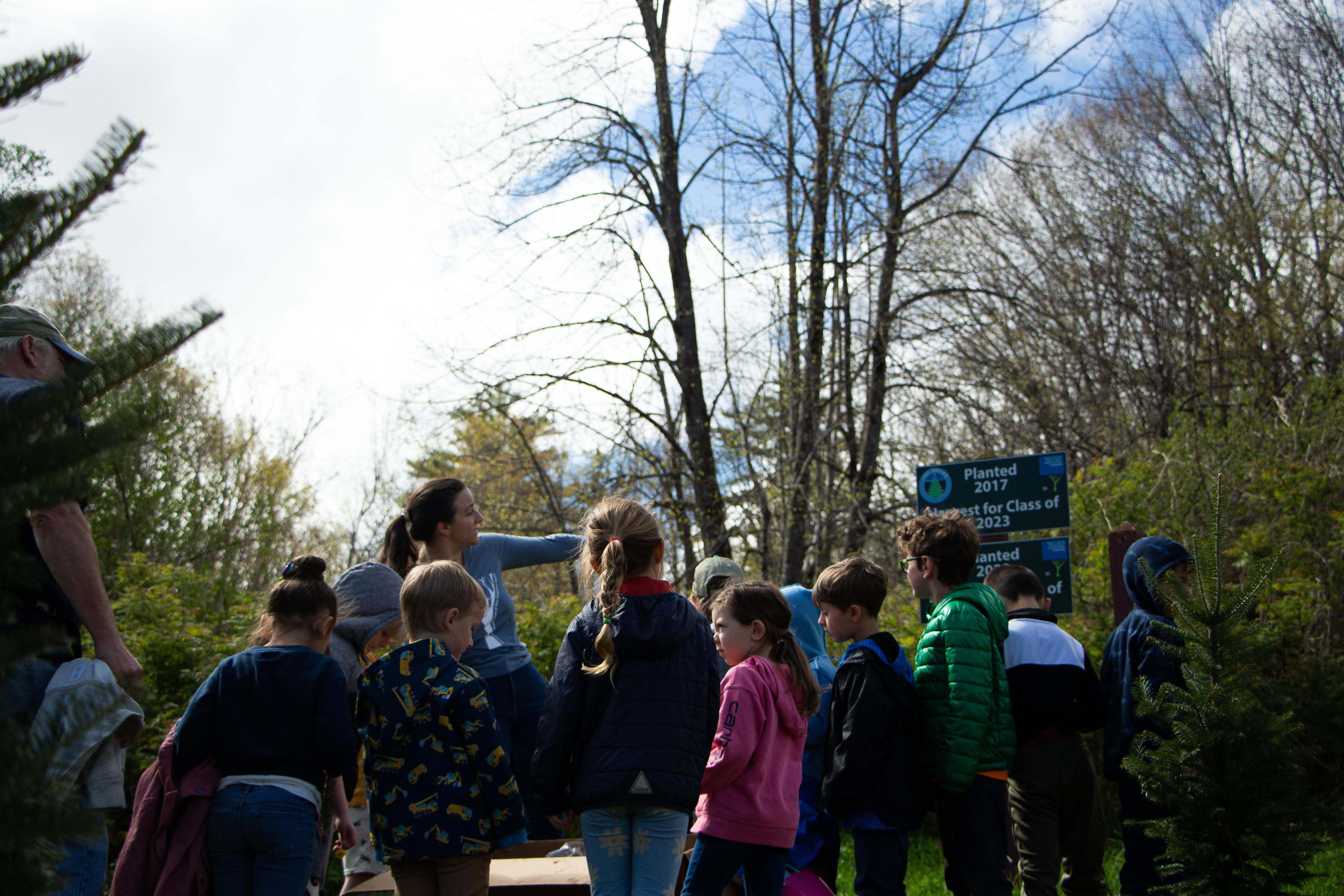 A group of children stand facing a woman pointing at a sign 