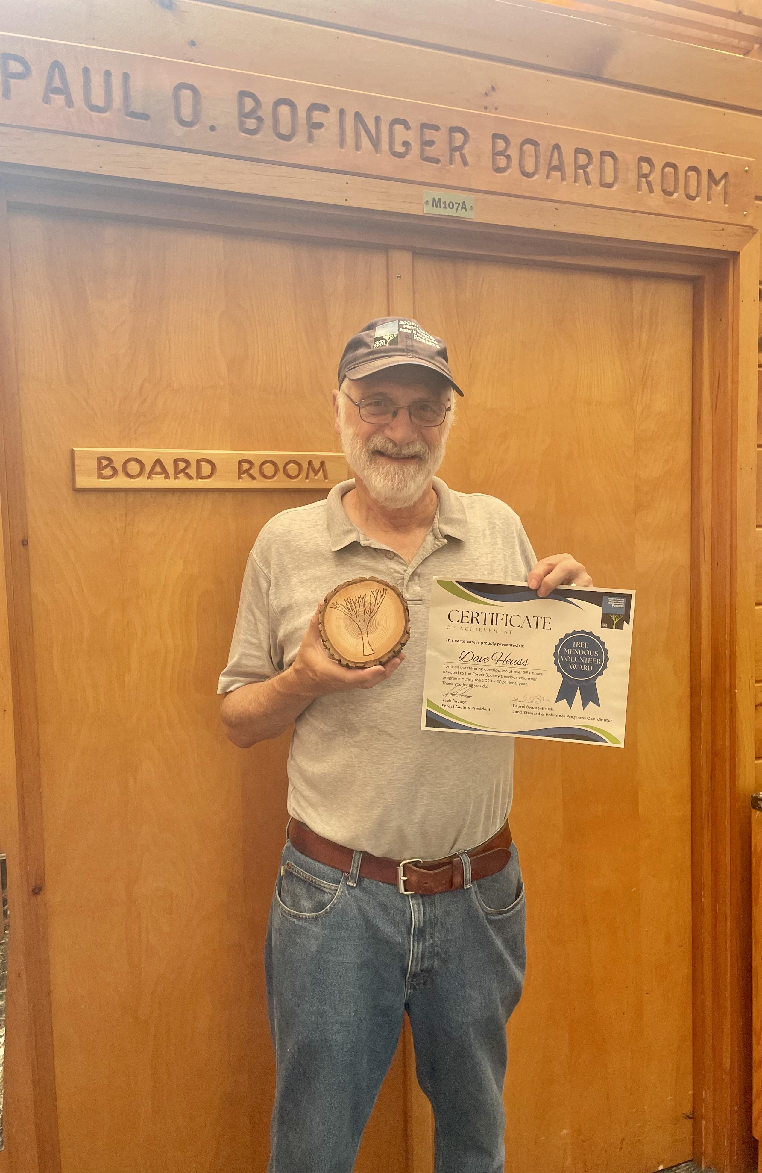 A man in a baseball hat holds up a paper certificate and a tree cookie award. 