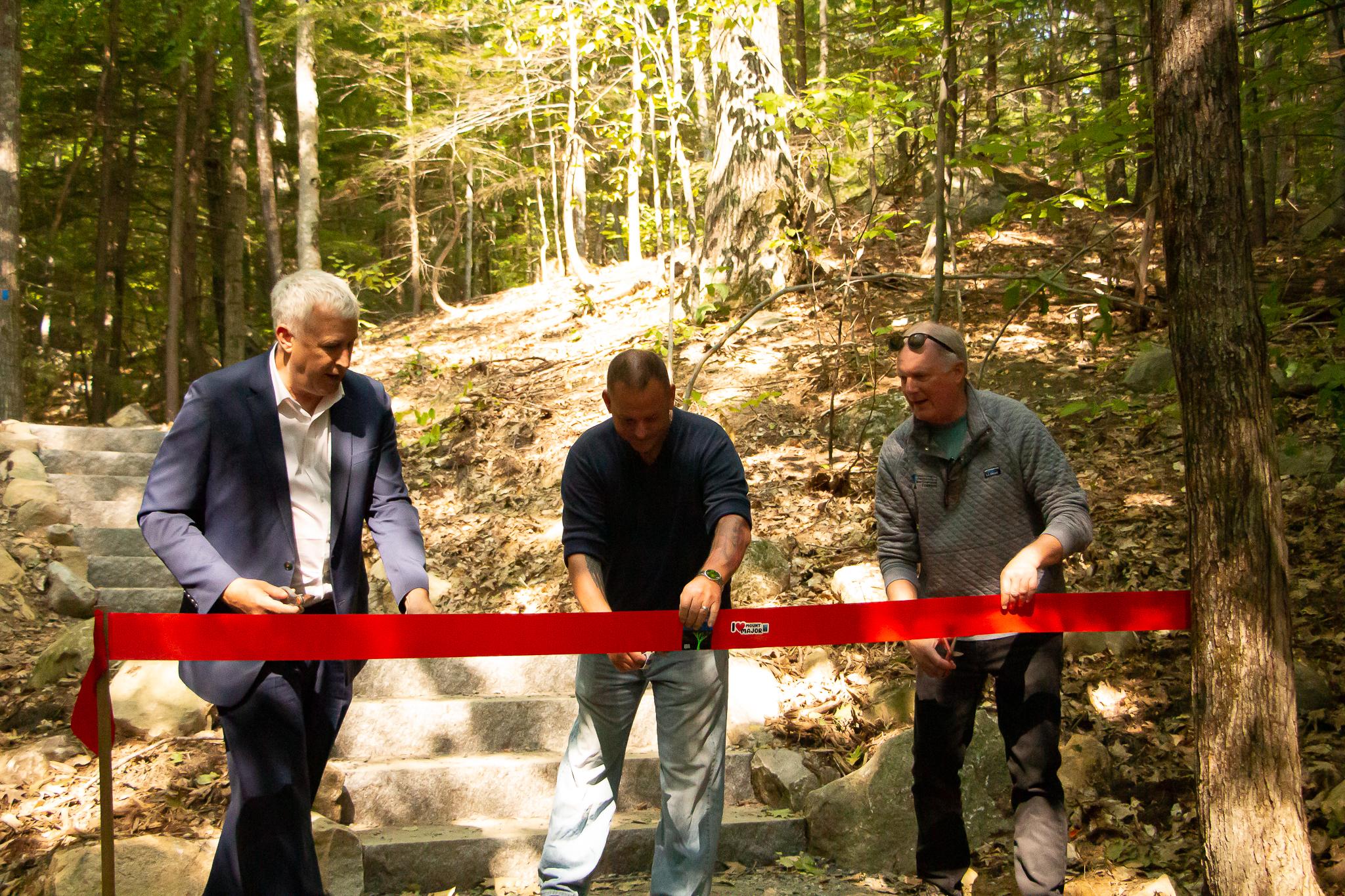 Three men cut a large red ribbon in front of a stone staircase on a trail in the woods