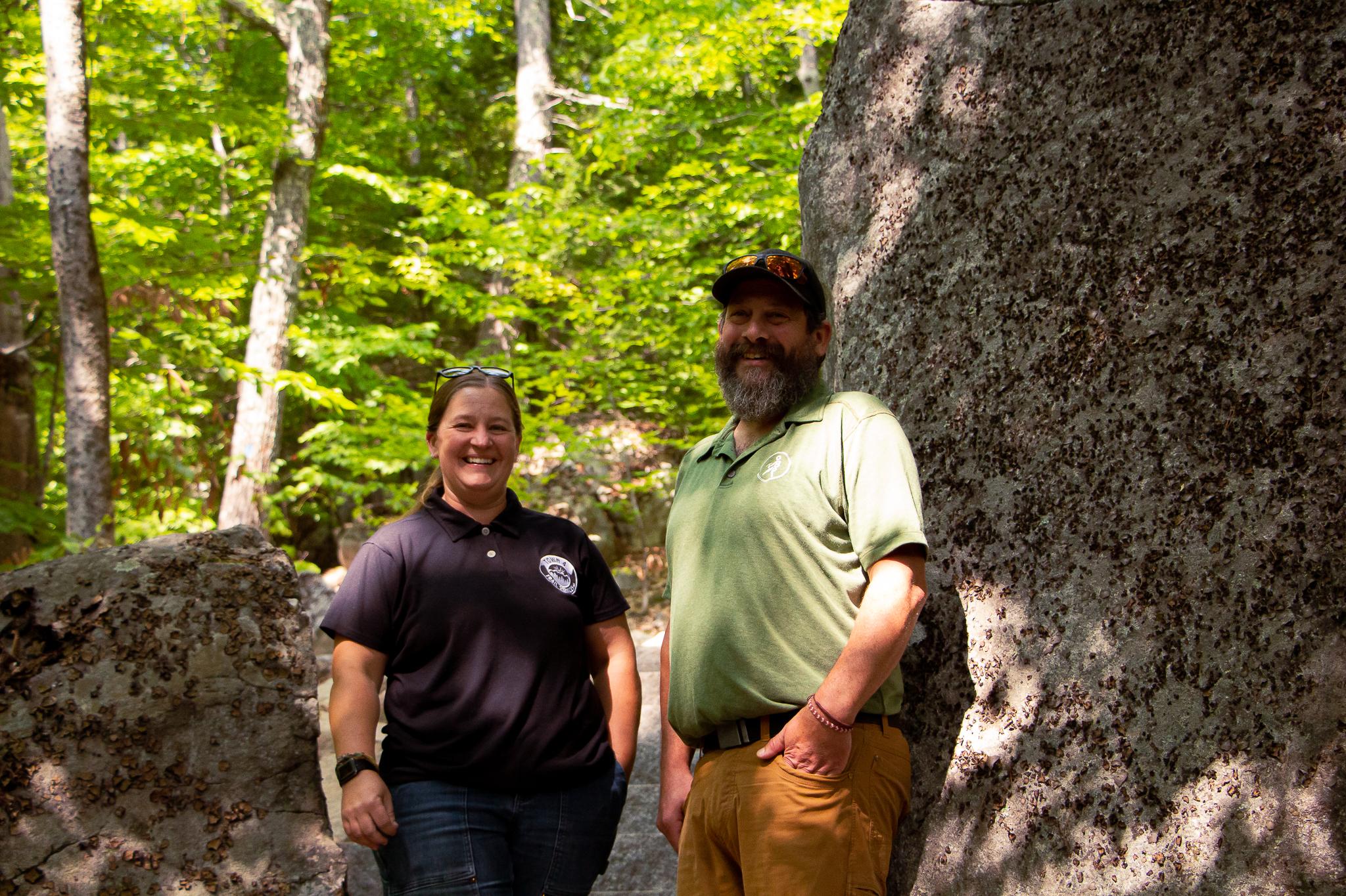 A man and a women stand together in front of a large boulder in the woods. 