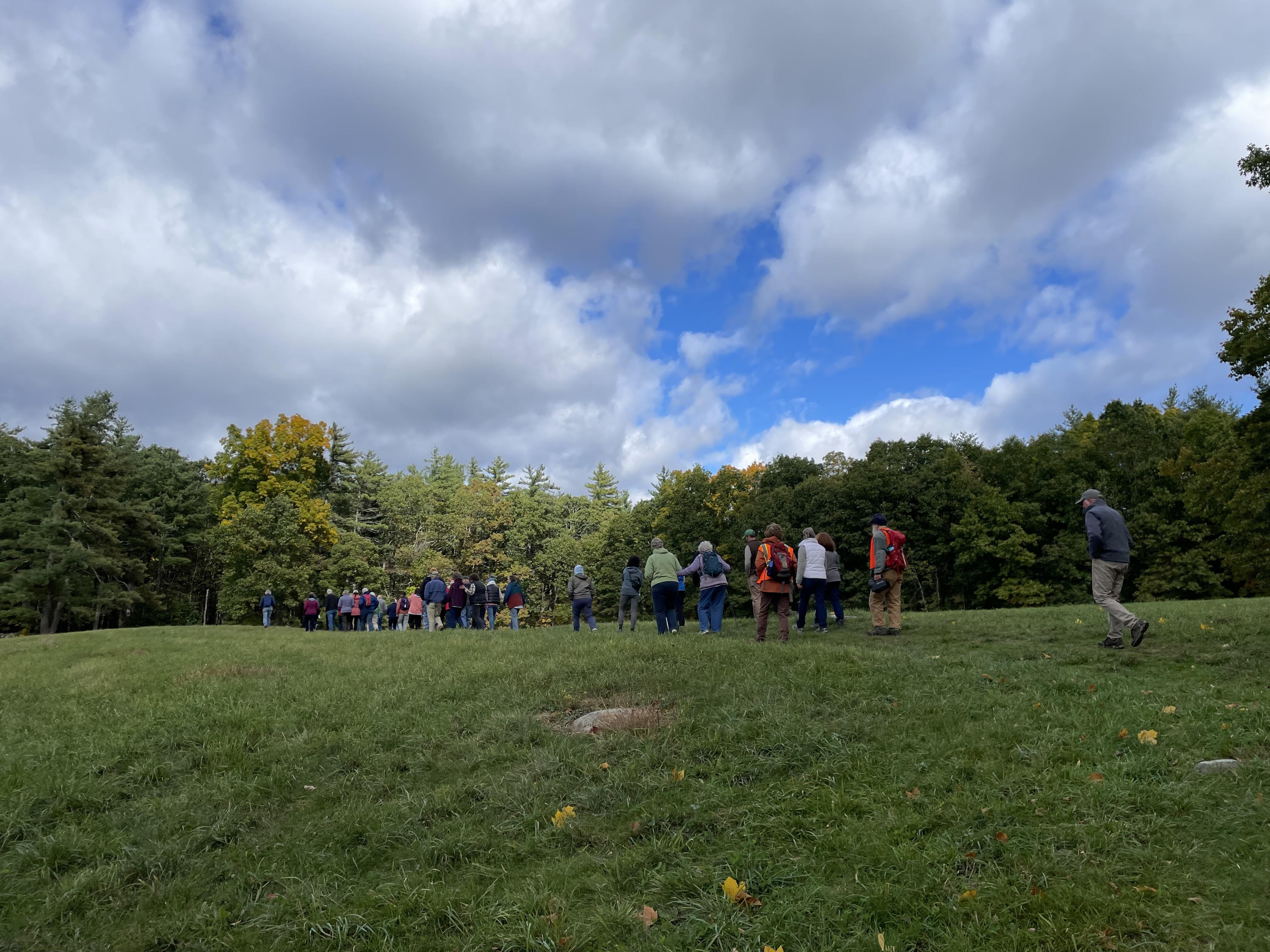 A line of hikers climb a small hill in a field towards a line of trees in the distance.