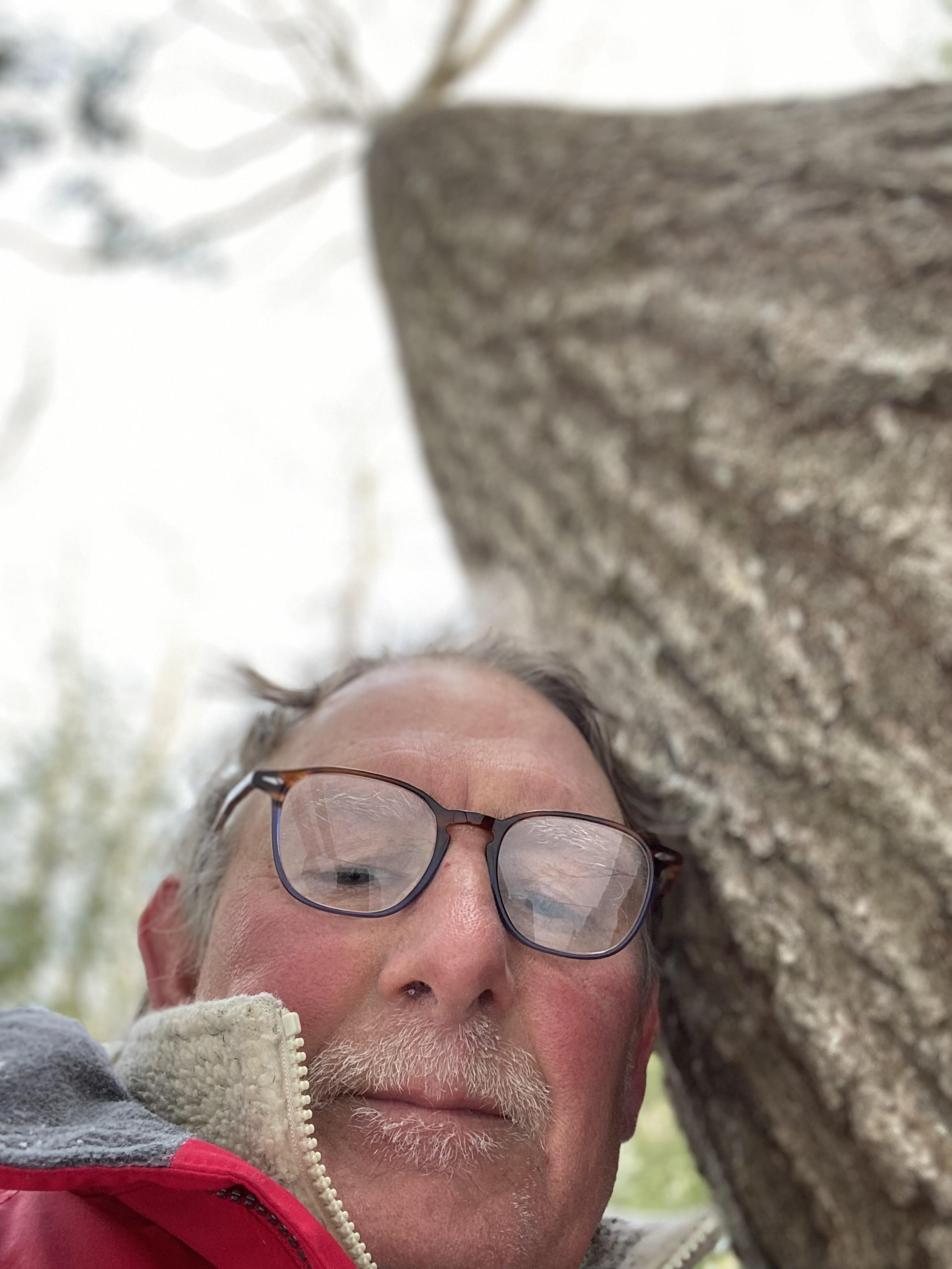 Man with glasses leans against a large tree trunk