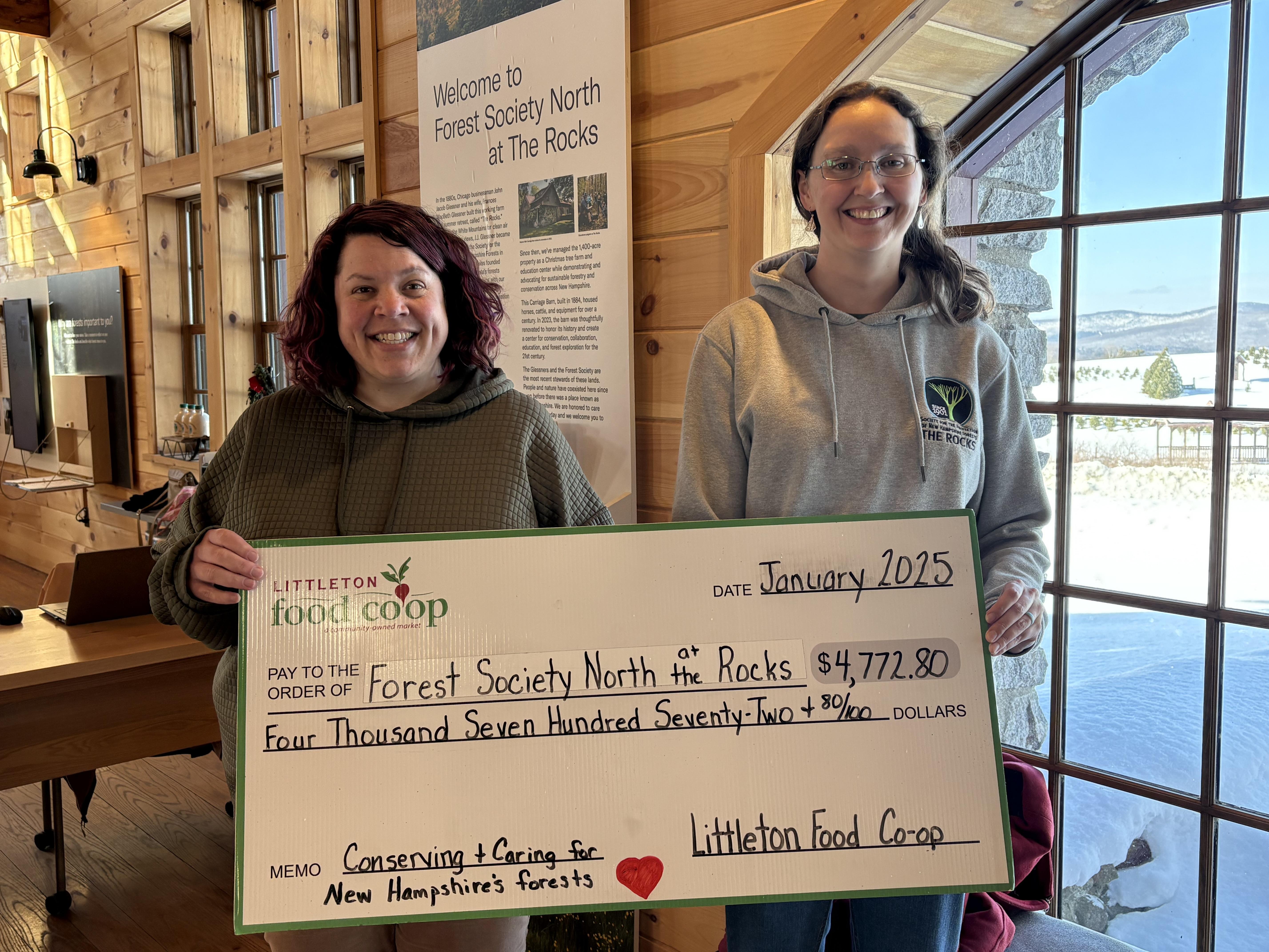 Two women stand in front of a large window holding a large donation check 