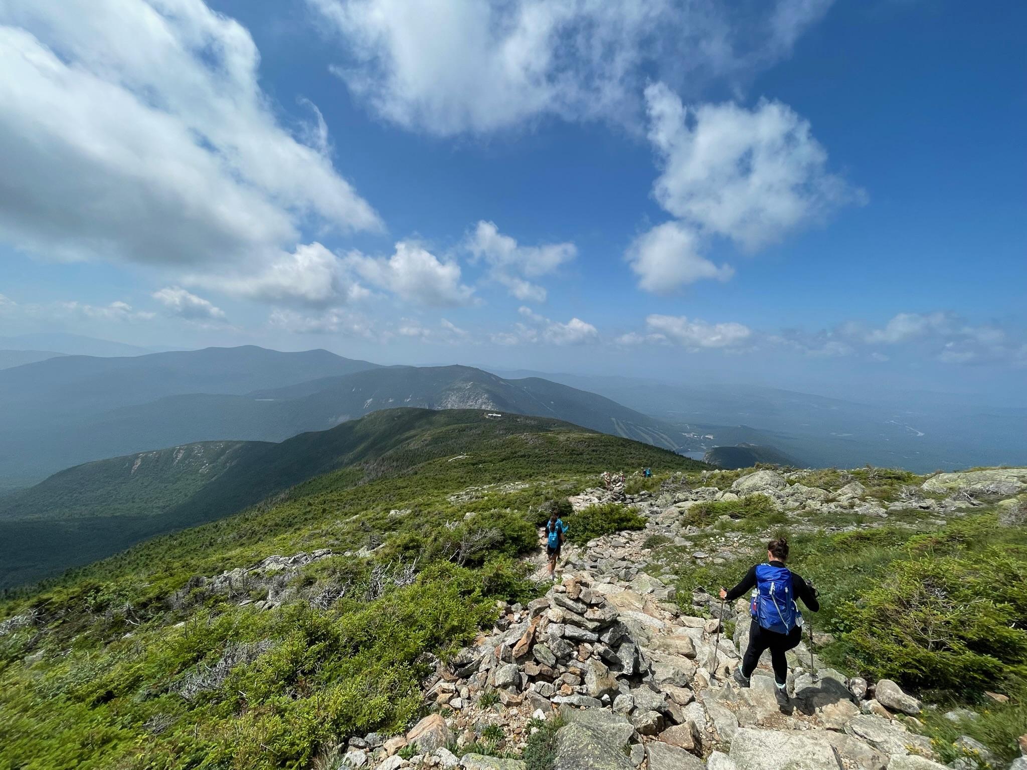 A woman with a blue backpack hikes over a rocky alpine trail. 