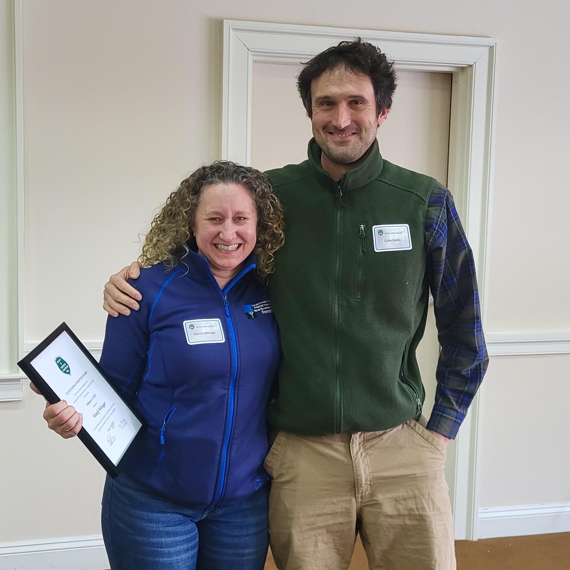 A man and woman stand side by side, the woman holds a medium sized plaque award