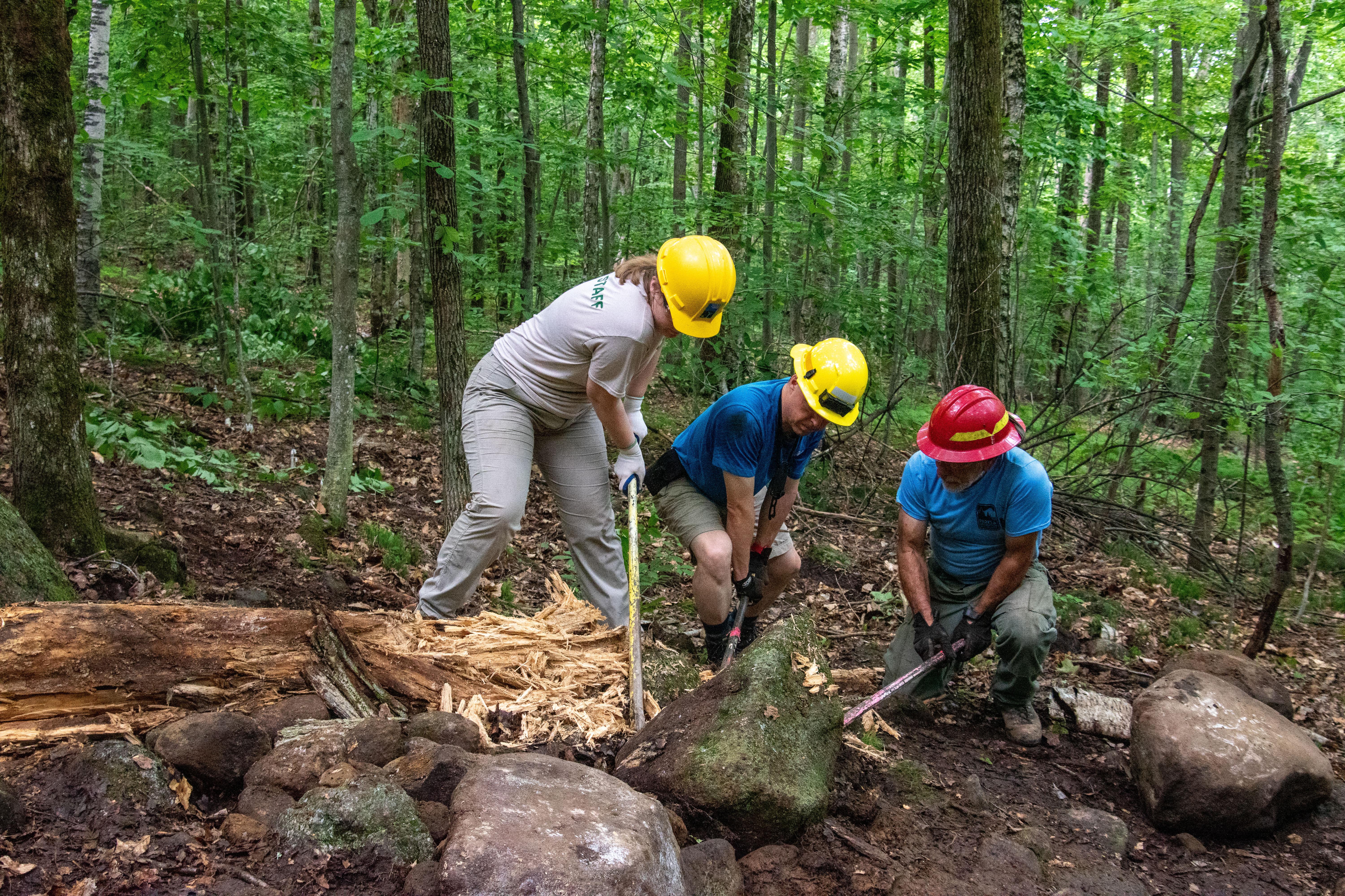 Two men and a woman wearing hard hats pry a large rock off the ground with rock bars. 