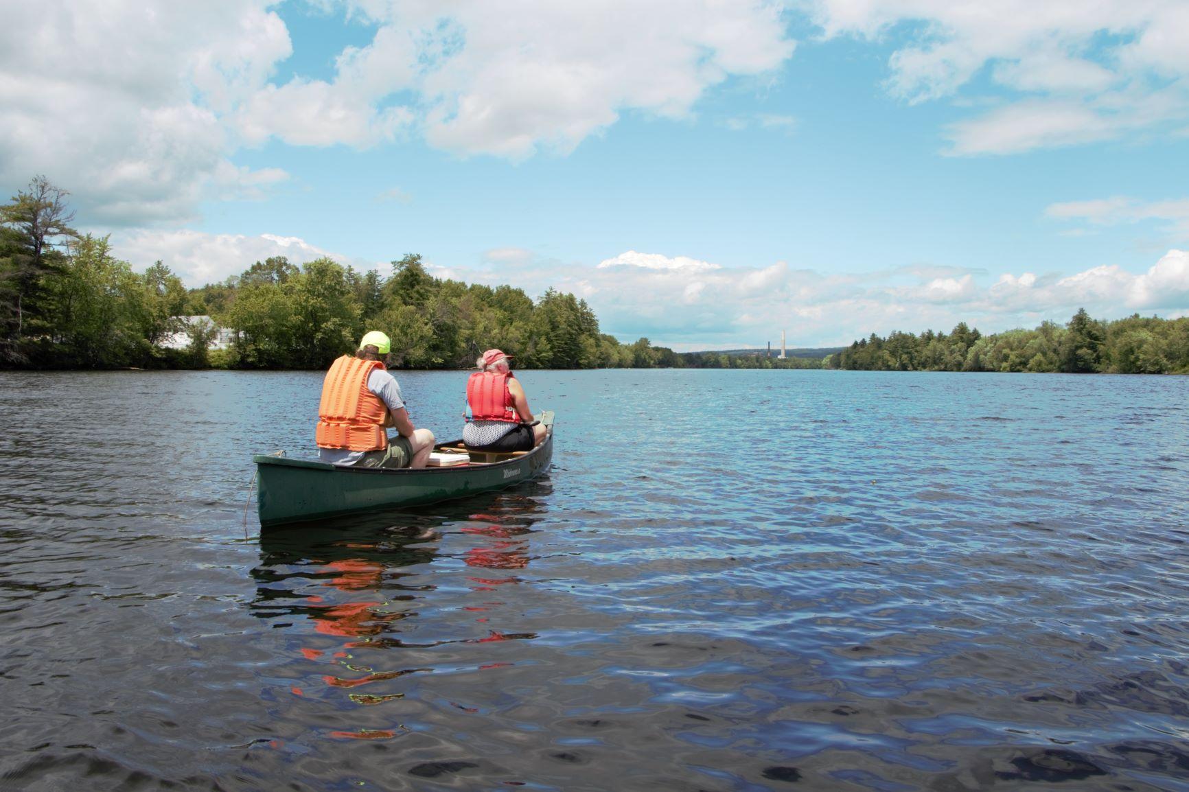 Two people in lifejackets in a canoe on the Merrimack River as seen from behind.