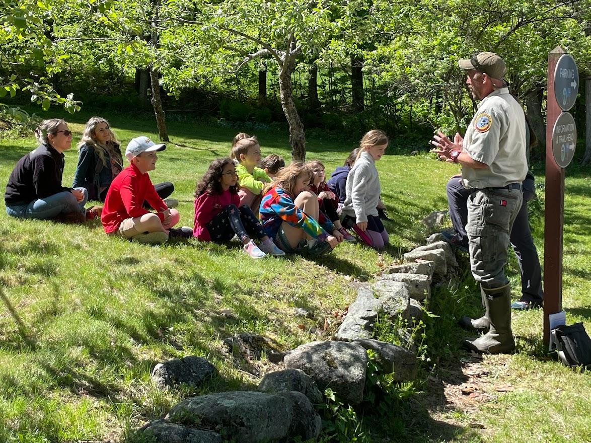 A group of school children sit on the lawn while a man speaks to them 