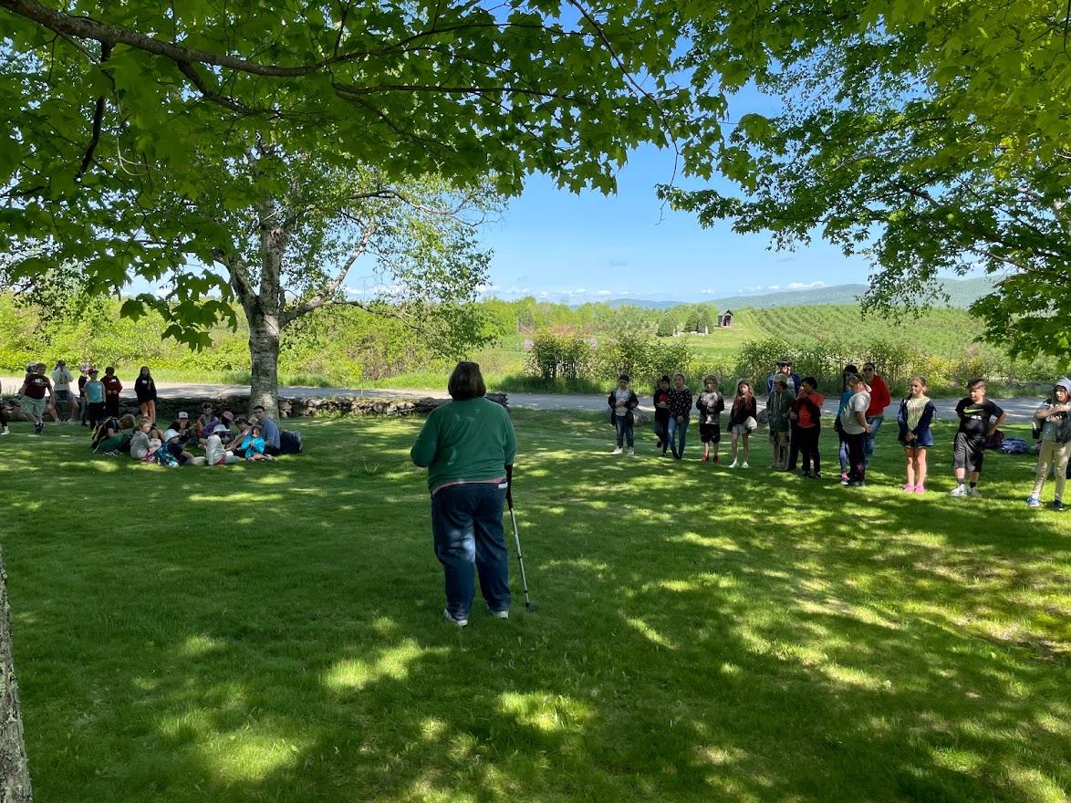 Three groups of fourth graders stand in a field while a woman with a cane speaks to them 