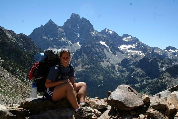 A woman wearing a backpack sits on a rock in front of a large mountain