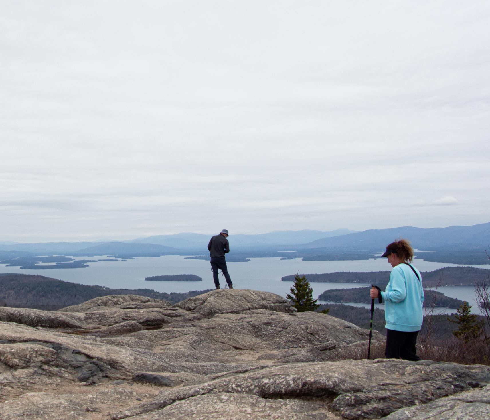 A woman in a blue shirt holding hiking poles hikes towards a man looking out on a mountain vista. 