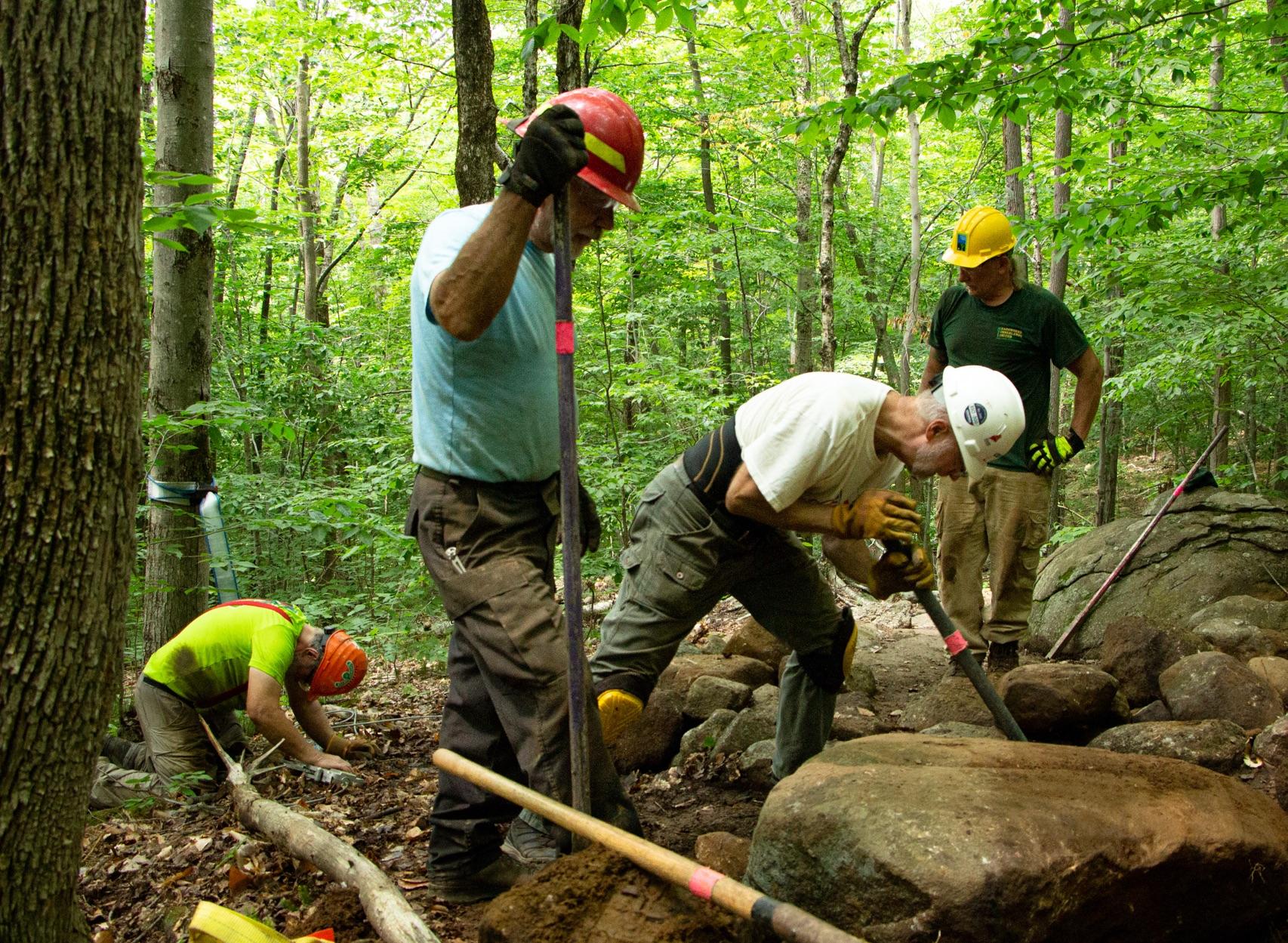 Three men wearing hardhats work to move a large rock. 