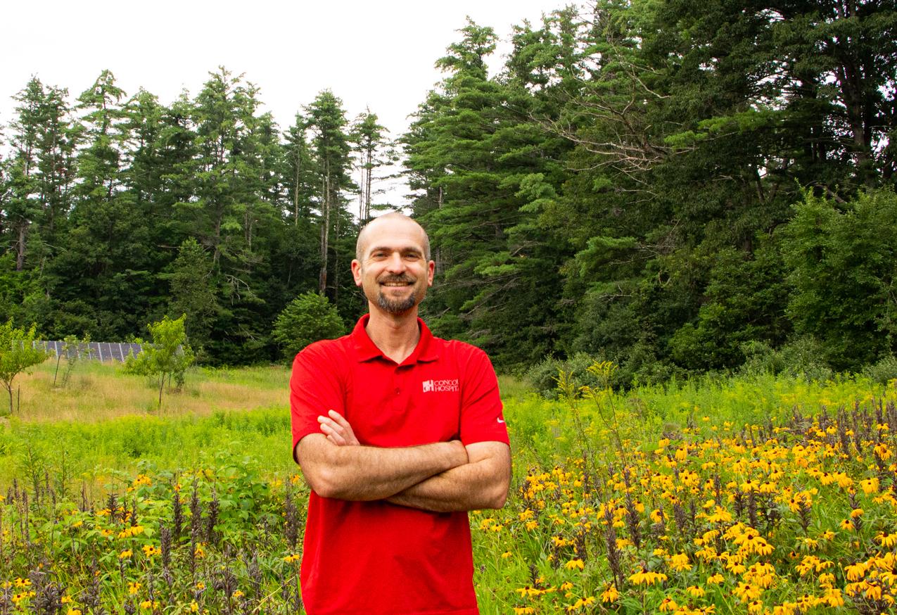 A man in a red shirt stands arms crossed in front of a large field 