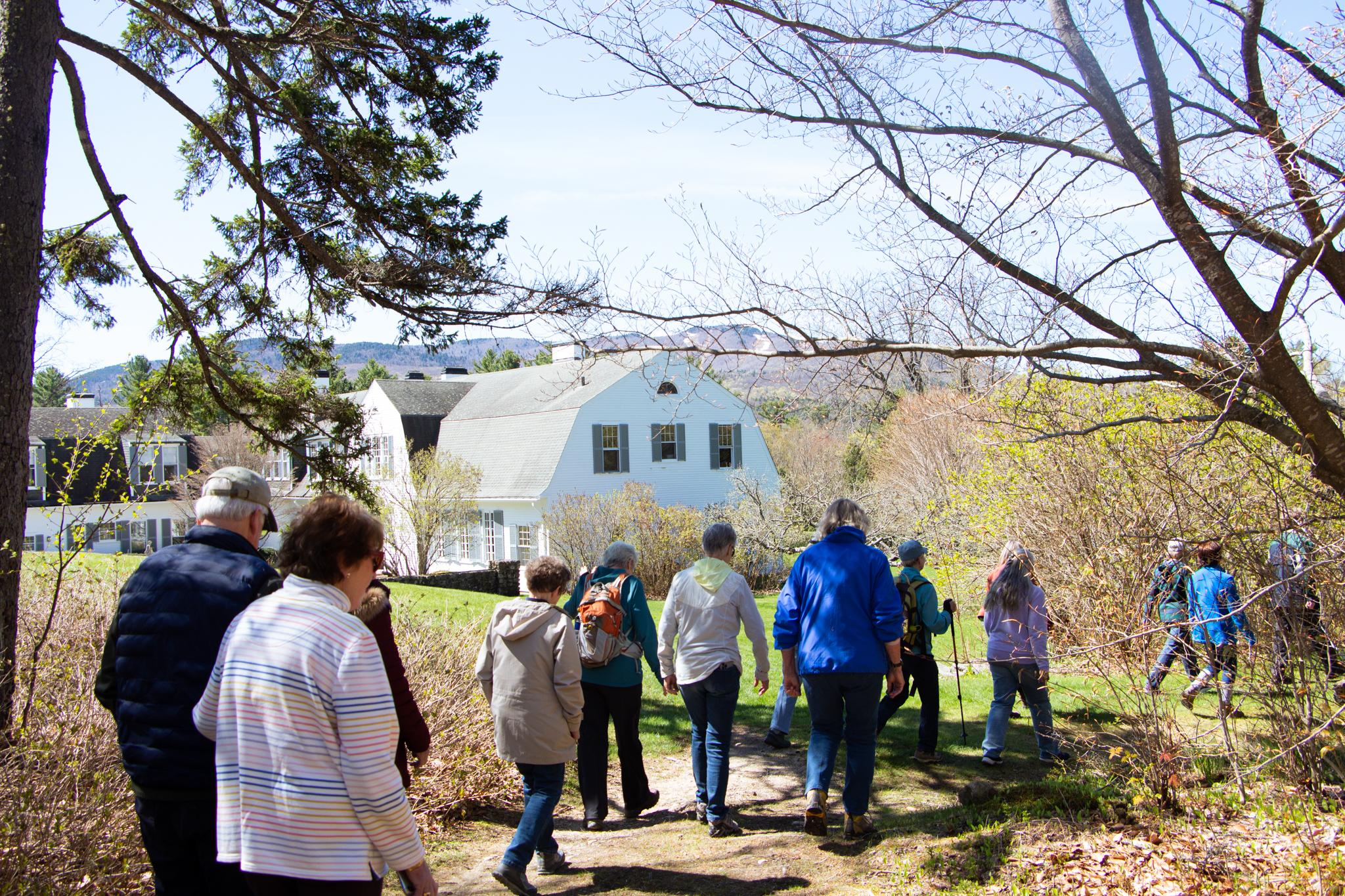 A group of hikers make their way down a trail, passed a large white house with green shutters. 