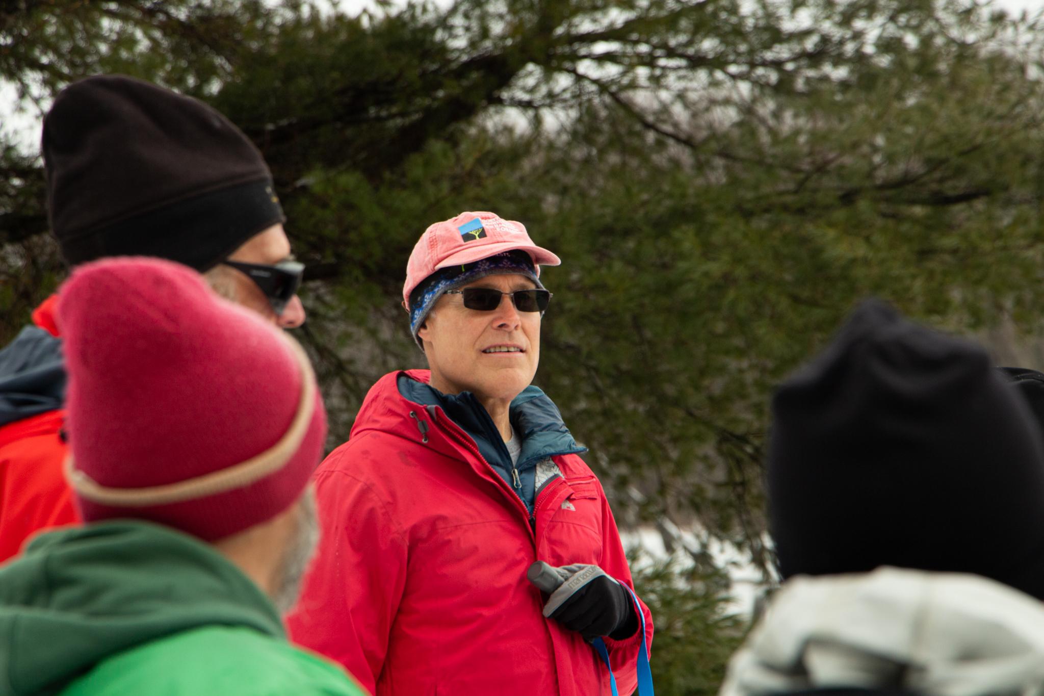 A man in a red jacket, pink baseball hat and sunglasses faces the camera amongst a group of people. 