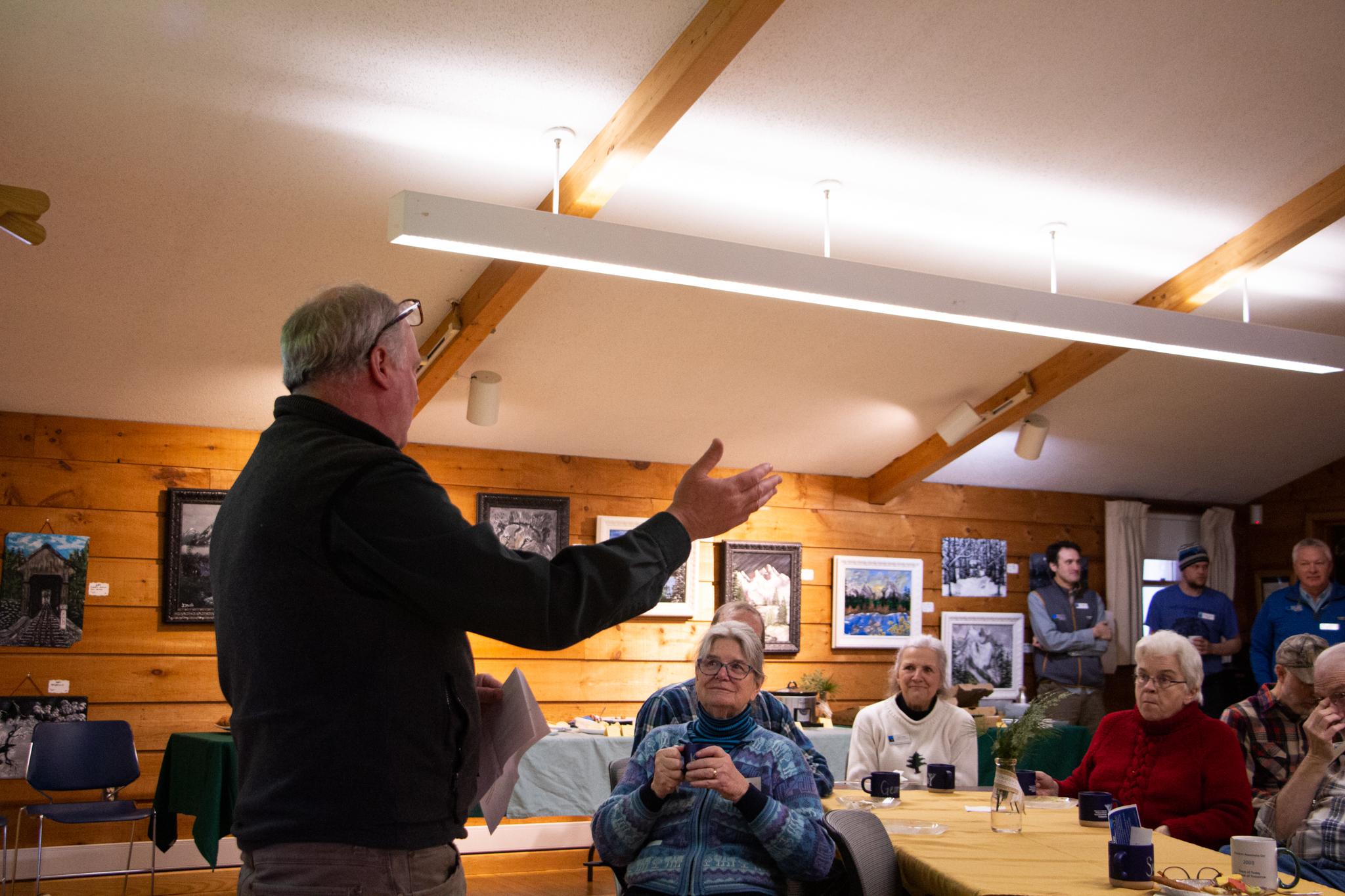 A man addresses a seated crowd inside a wood paneled room 