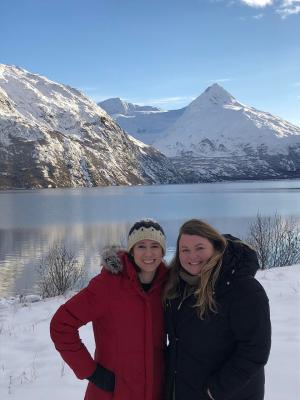 Two women in heavy coats stand in front of a snowy mountain 