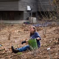 A woman sits in a garden, holding a green bucket. 