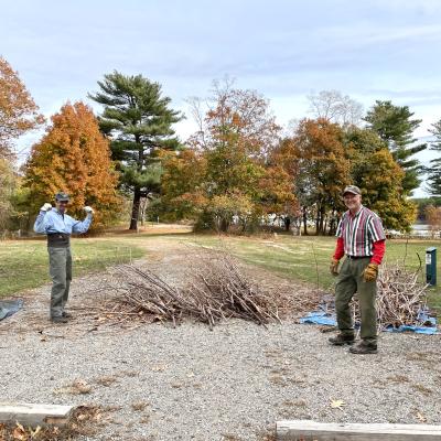 Two men stand in front of a large pile of brush. 