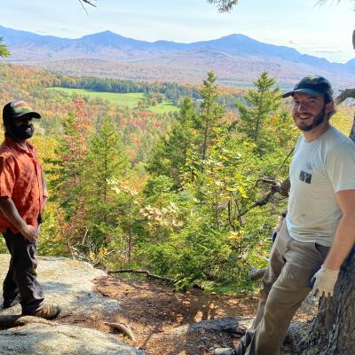 Two men stand on a rock overlooking a mountain view. 