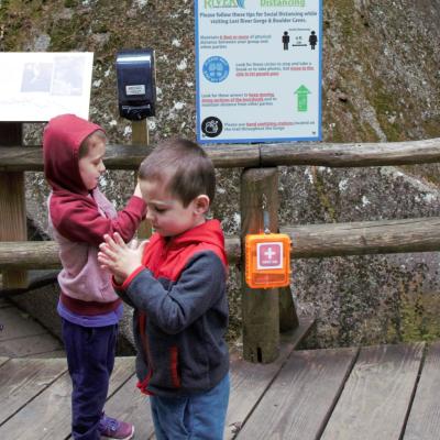 Two preschoolers wipe hands on a boardwalk near a hand sanitizing station.