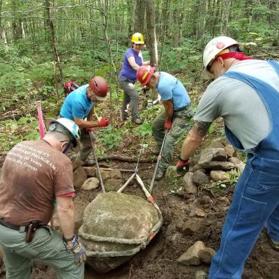 Volunteers in hard hats help lift a boulder from a trail.