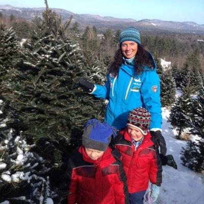 A woman and two children pose with a Christmas tree amid rows of snow-covered Christmas trees.