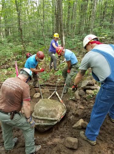 Volunteers in hard hats help lift a boulder from a trail.
