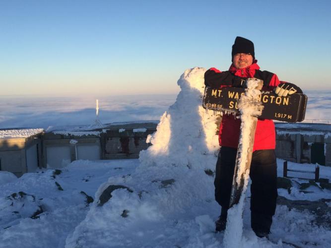 Dan Szczesny in red parka poses at rime ice crusted summit sign on Mt Washington in winter