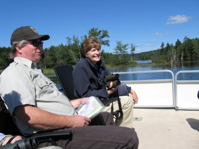 Senator Jeanne Shaheen smiling with binoculars sitting on boat with USFW Staff on Lake Umbagog on a clear blue sky day.
