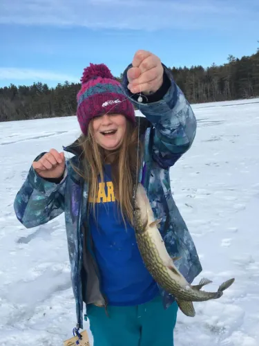 A young girl in winter clothes holds up a long fish on a fishing line 