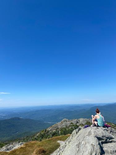 Woman sits on the edge of a large rock at the top of a mountain looking out to the view 