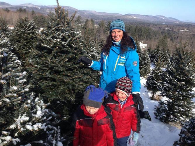 A woman and two children pose with a Christmas tree amid rows of snow-covered Christmas trees.