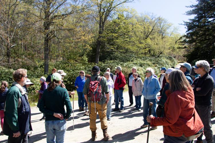 A man in a red backpack addresses a large crowd of people dressed for hiking. 