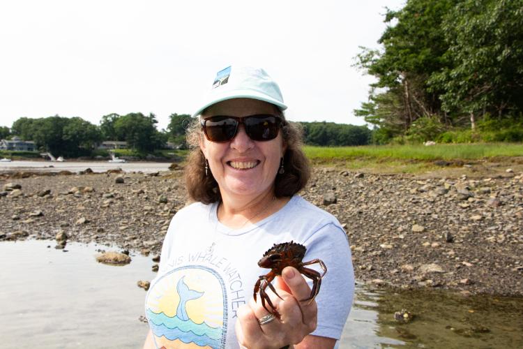 A woman wearing a baseball hat and sunglasses holds a crab out to the camera. 