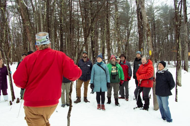 A man in a red jacket faces away from camera as he speaks to a crowd in a snowy forest. 